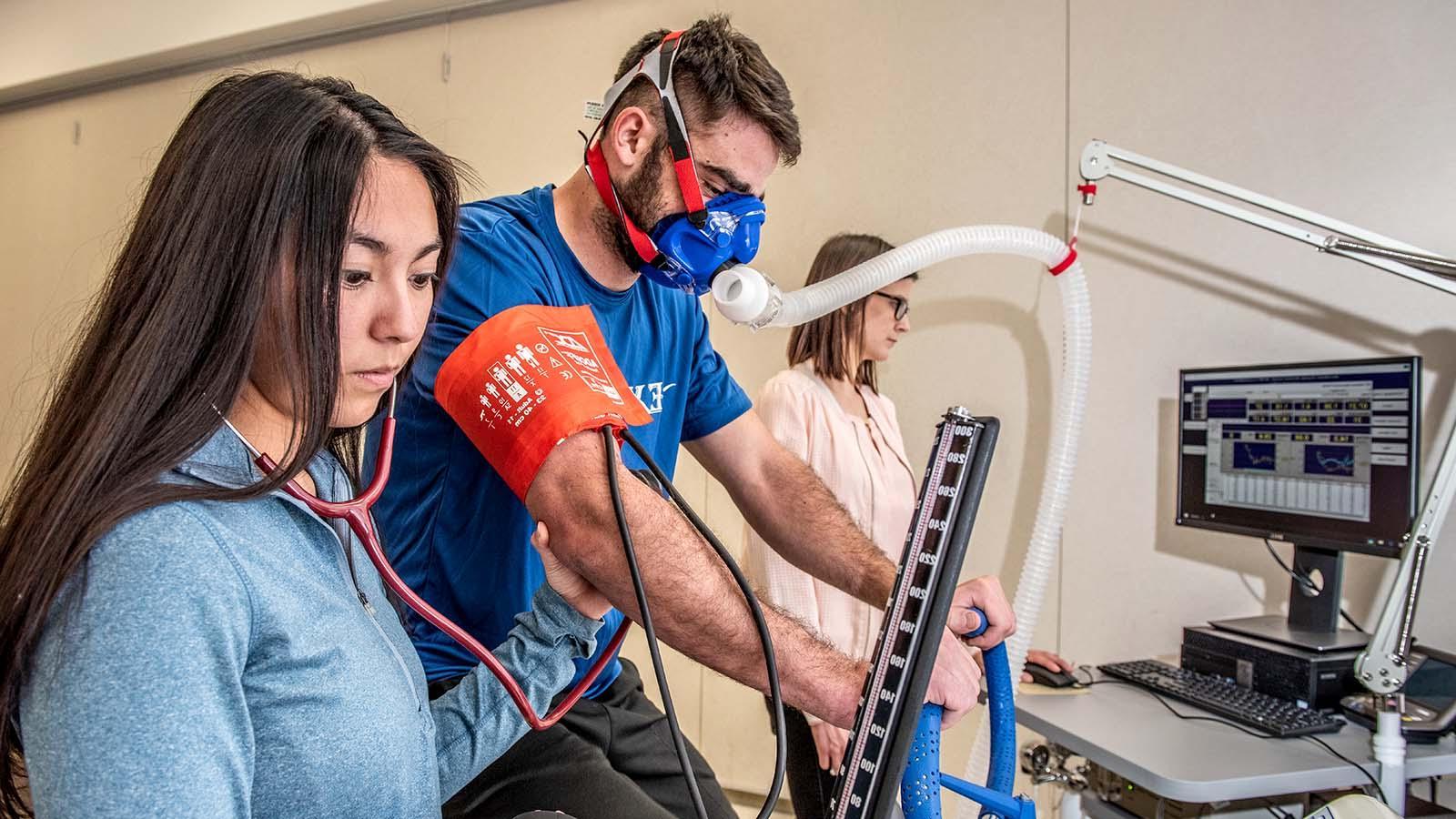 exercise science student monitoring the breathing of an athlete riding an exercise bike and wearing a VO2 max mask.